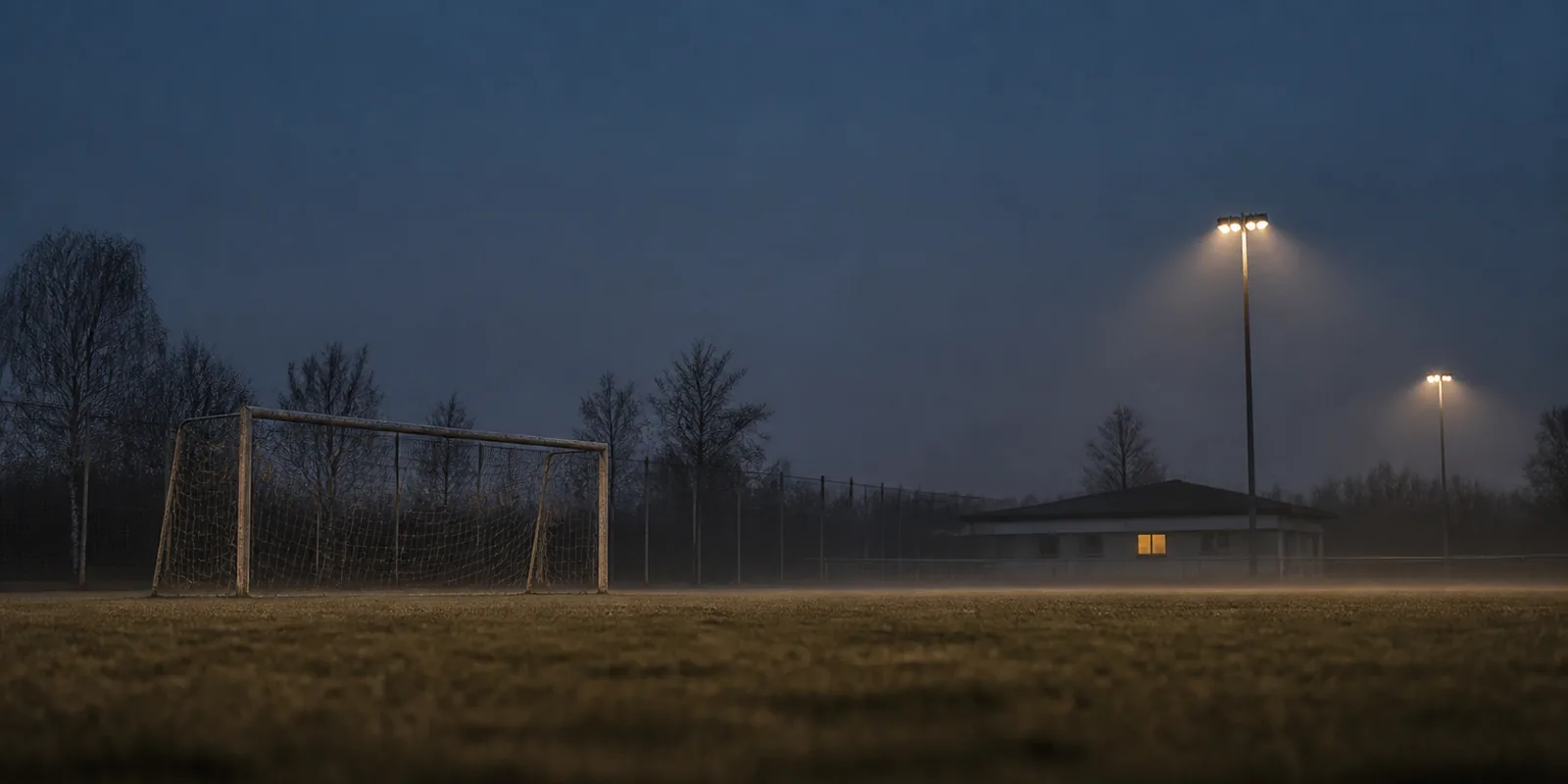 Ein leerer Amateurfußballplatz in der Abenddämmerung. Ein verwittertes Tor mit durchhängendem Netz, dahinter ein Zaun, kahle Bäume und ein niedriges Vereinsheim mit einem einzigen erleuchteten Fenster. Zwei Flutlichtmasten leuchten in den Bodennebel.